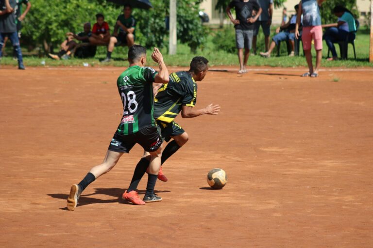 Two athletes engage in a competitive soccer match on a dirt field with an audience.